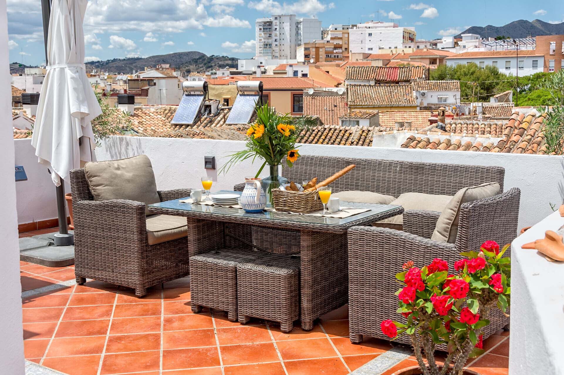 A terrace with wicker furniture, a table set for breakfast, and a view of the cityscape.