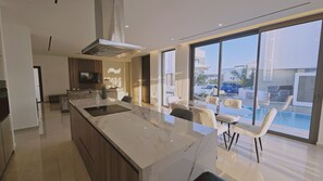 Une cuisine moderne dotée d’un grand îlot central, d’un coin repas avec des chaises blanches et d’une vue sur une piscine et des bâtiments.