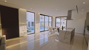 A modern kitchen with a marble countertop, a dining area with a glass table, and large windows offering a view of a swimming pool and buildings. - STAY Oasis Luxury Villa 5 (Ayia Napa)