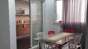 A dining area with a glass table and white chairs, a kitchen in the background, and a window with curtains. - Luxueux Appartement S+3 Avec Jacuzzi - Confort et Élégance (nabeul)