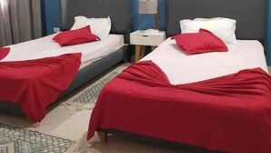 A hotel room with two beds, each with a red bedspread and white pillows. The room has a blue accent wall with hexagonal wall art. There is a small bedside table with a lamp and a white box on it. - Luxueux Appartement S+3 Avec Jacuzzi - Confort et Élégance (nabeul)