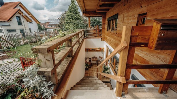 Un escalier en bois mène à un balcon situé au deuxième étage, d’où l’on peut admirer un jardin et les maisons voisines.