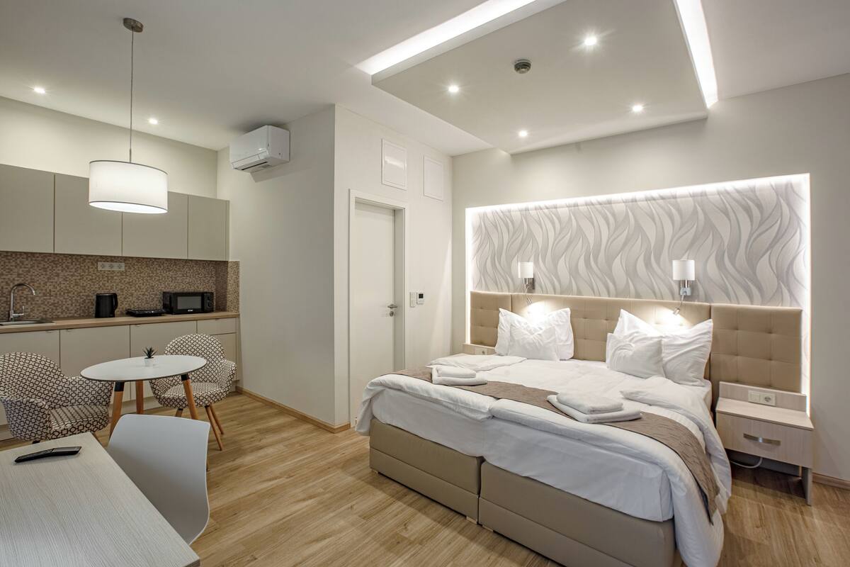 A modern hotel room with a large bed, a small dining table, and a kitchenette.