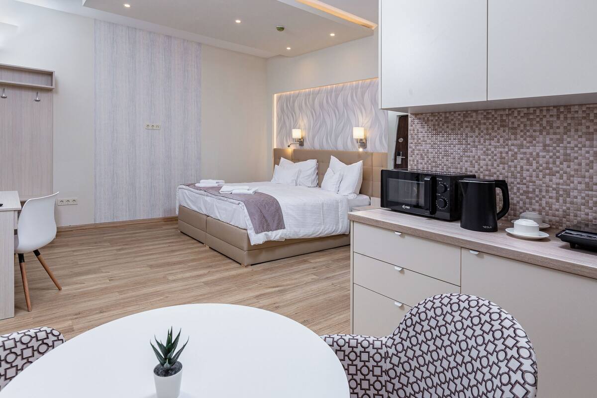 A modern hotel room with a bed, a desk, a chair, a dining table, and a kitchenette.