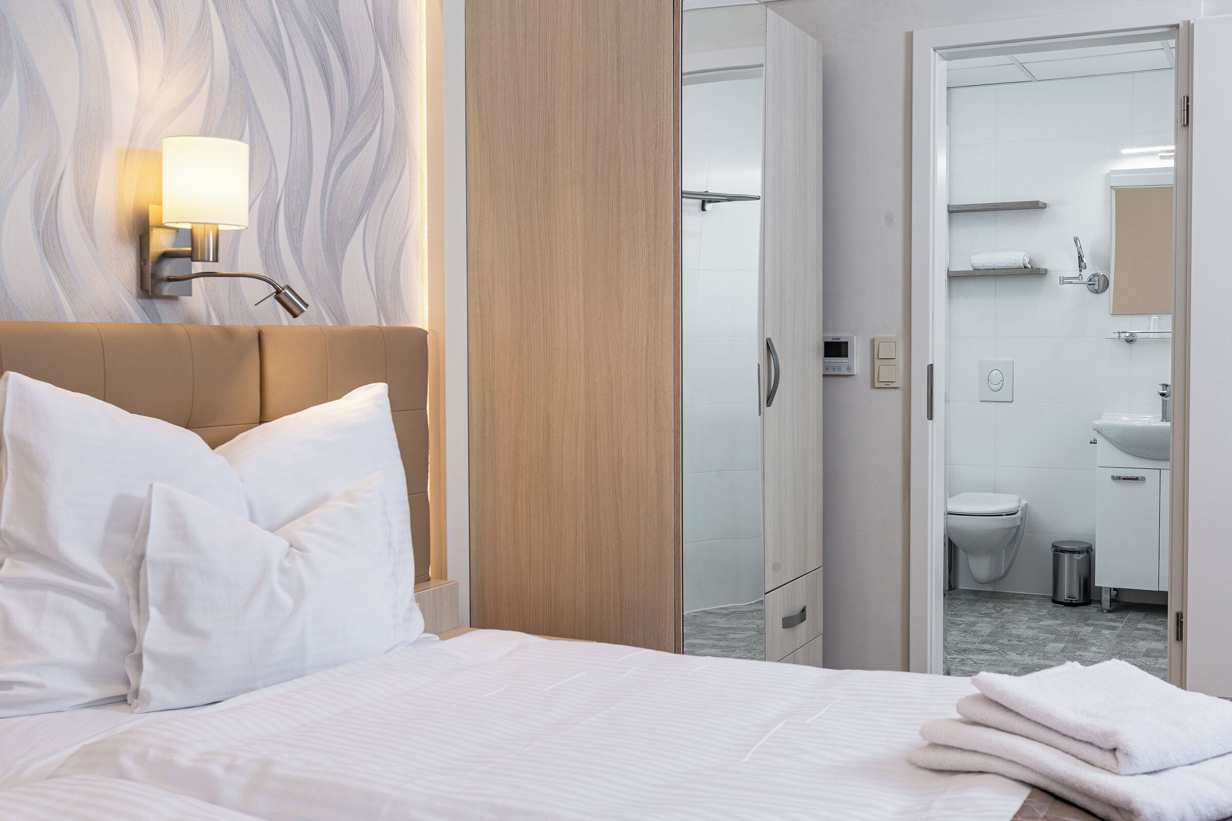 Ein Hotelzimmer mit einem Bett, einem Nachttisch, einer Wandleuchte, einem Holzschrank und einem durch eine offene Tür einsehbaren Badezimmer.