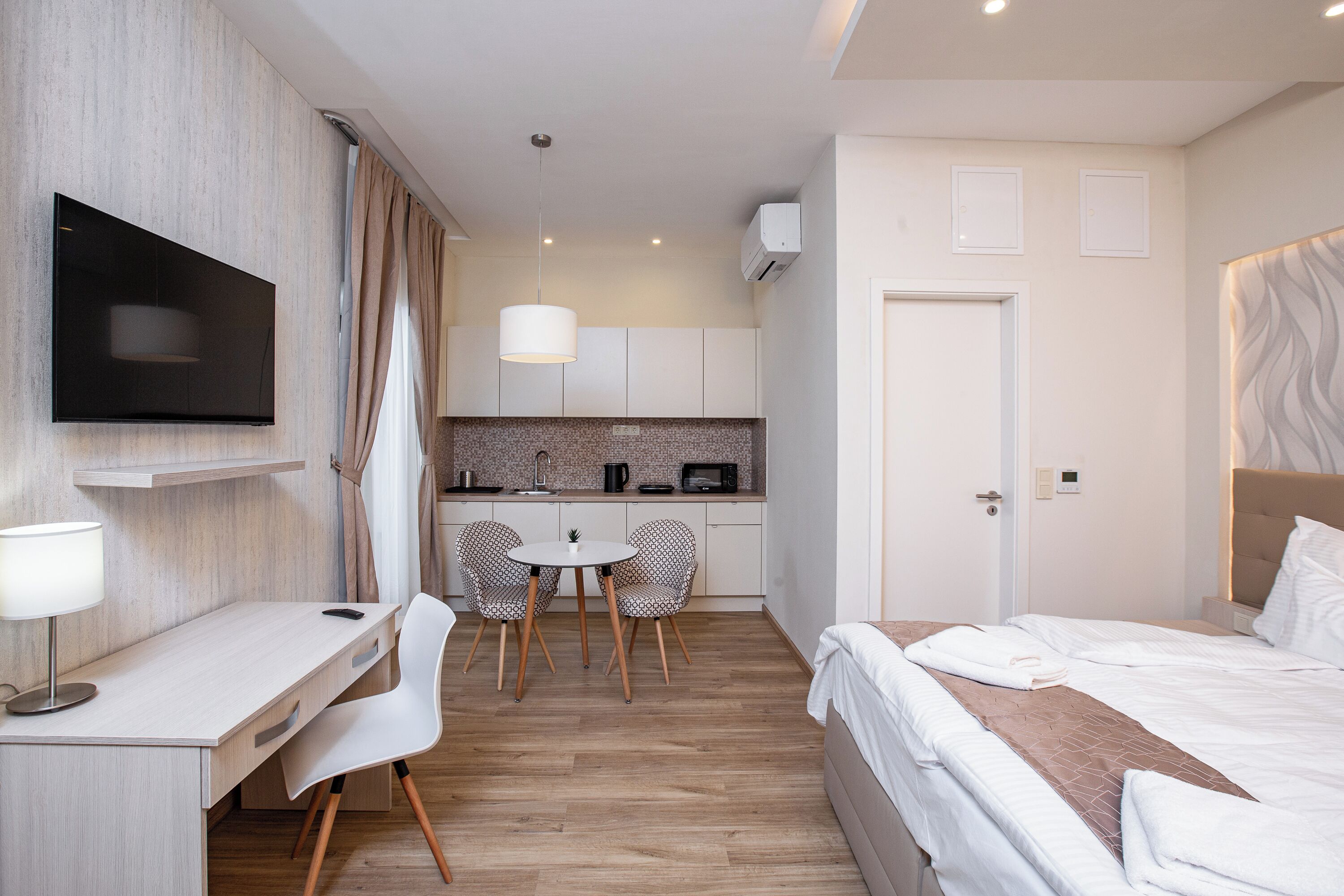 A hotel room with a bed, a desk, a TV, a small kitchen area, and a dining table with chairs.
