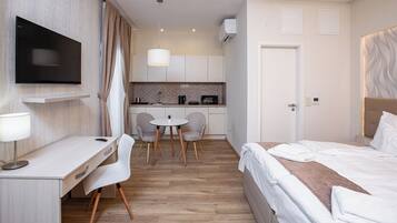 A hotel room with a bed, a desk, a TV, a small kitchen area, and a dining table with chairs.