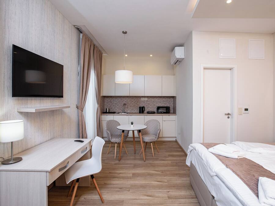 A hotel room with a bed, a desk, a TV, a small kitchen area, and a dining table with chairs.