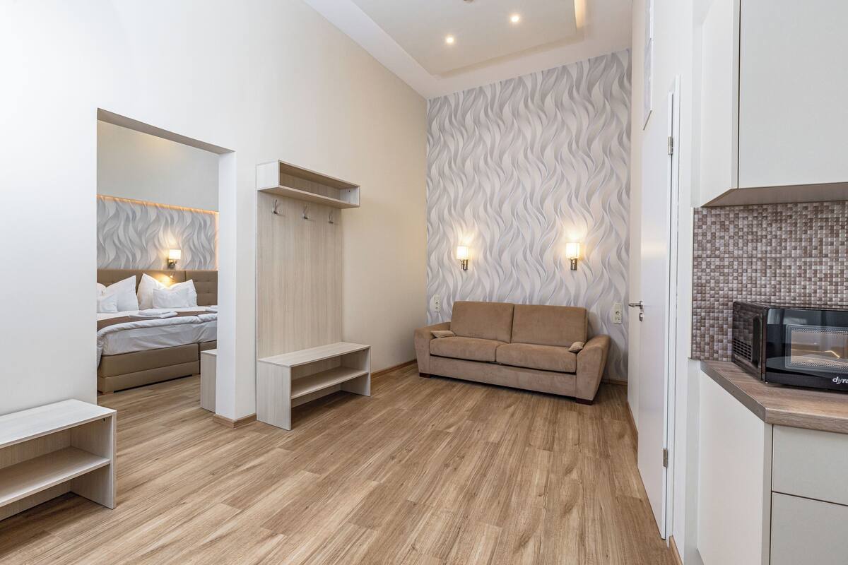 A modern bedroom with a bed, bedside table, and a built-in bench. There is a sofa and a microwave in the adjacent room.