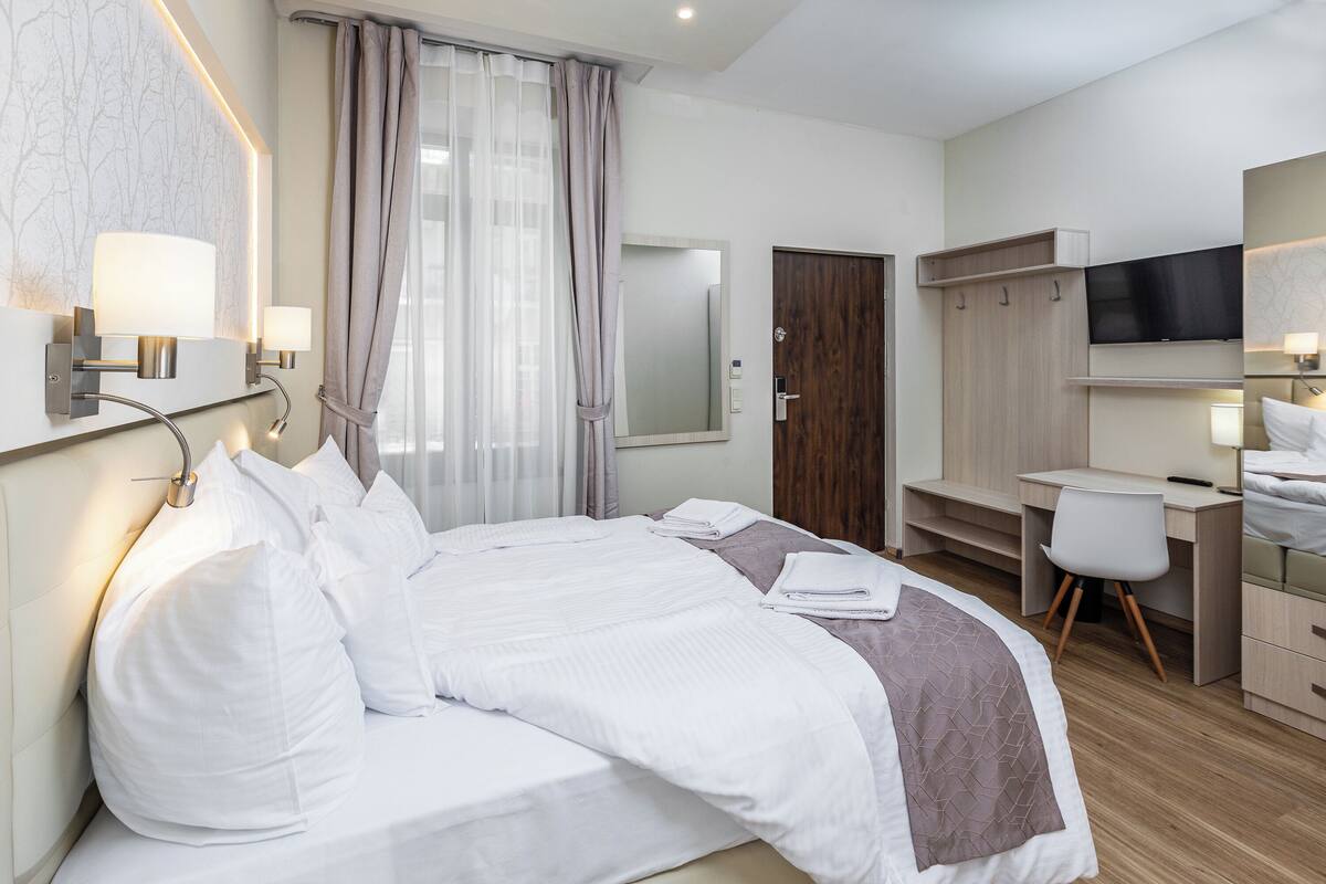 A modern hotel room with a neatly made bed, a desk with a chair, a TV, and a wardrobe.