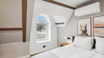 A bedroom with a large bed, a nightstand, a lamp, a window with a view of snow-covered trees, and two framed pictures on the wall.