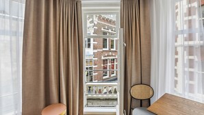 A room with a view of a brick building, a small round table, a bench, and a window with curtains. - Limehome Den Haag Nieuwstraat (The Hague)