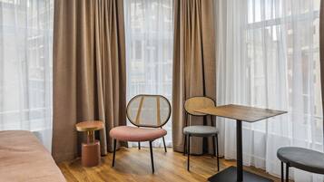 A modern hotel room with a large bed, a small round table, two chairs, and a view through sheer curtains.