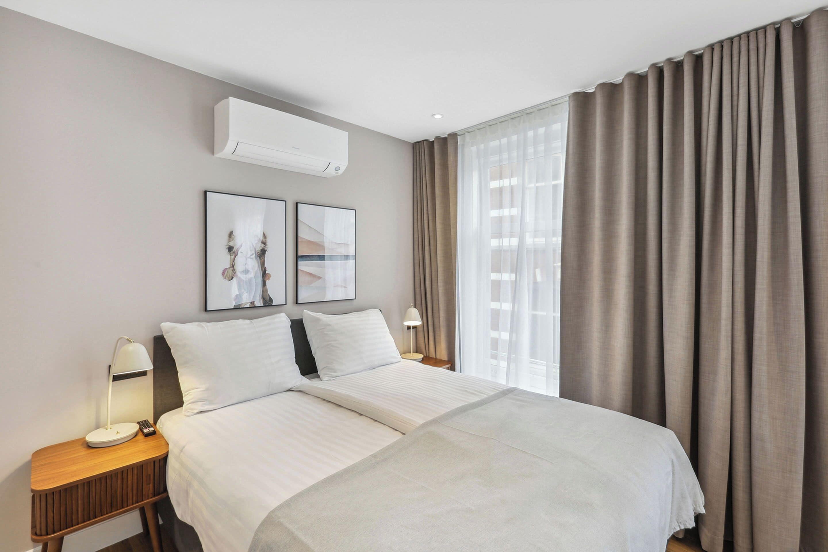 A hotel room with a large bed, a nightstand with a lamp, an air conditioning unit, and a window with curtains.