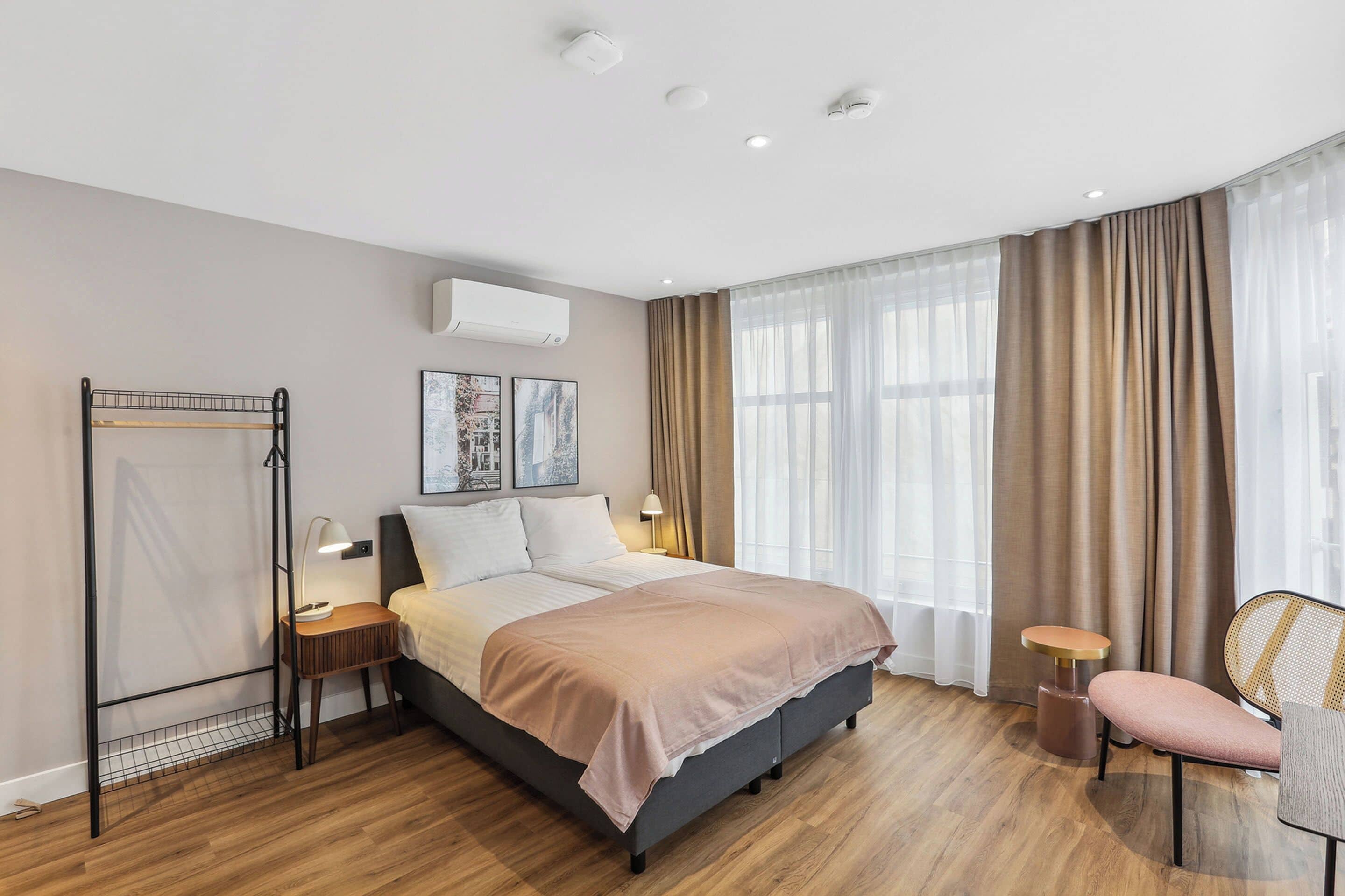 A modern hotel room with a large bed, a bedside table with a lamp, a chair, and a window with curtains.