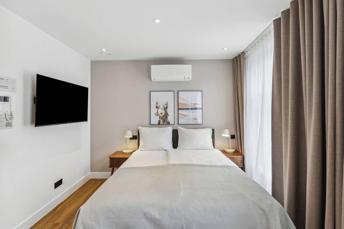 A modern hotel room with a large bed, a flat-screen TV mounted on the wall, an air conditioning unit, and two bedside tables with lamps.