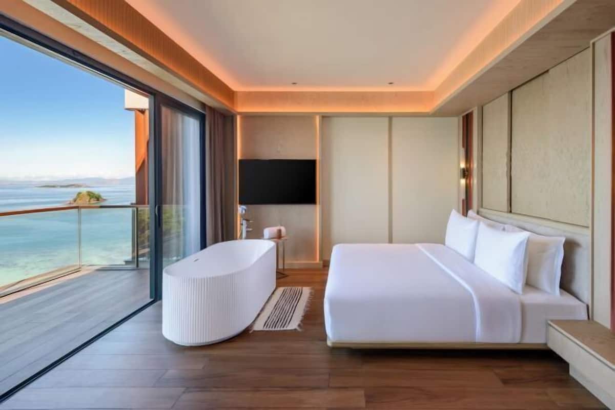 Ein modernes Hotelzimmer mit einem großen Bett, einer Badewanne, einem Flachbildfernseher und einem Balkon mit Meerblick.