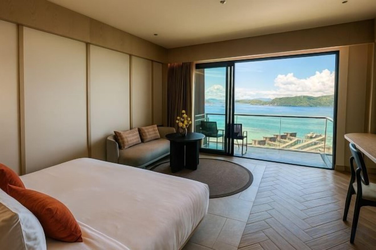 A hotel room with a bed, a sofa, a small table, and a balcony with a view of the sea.