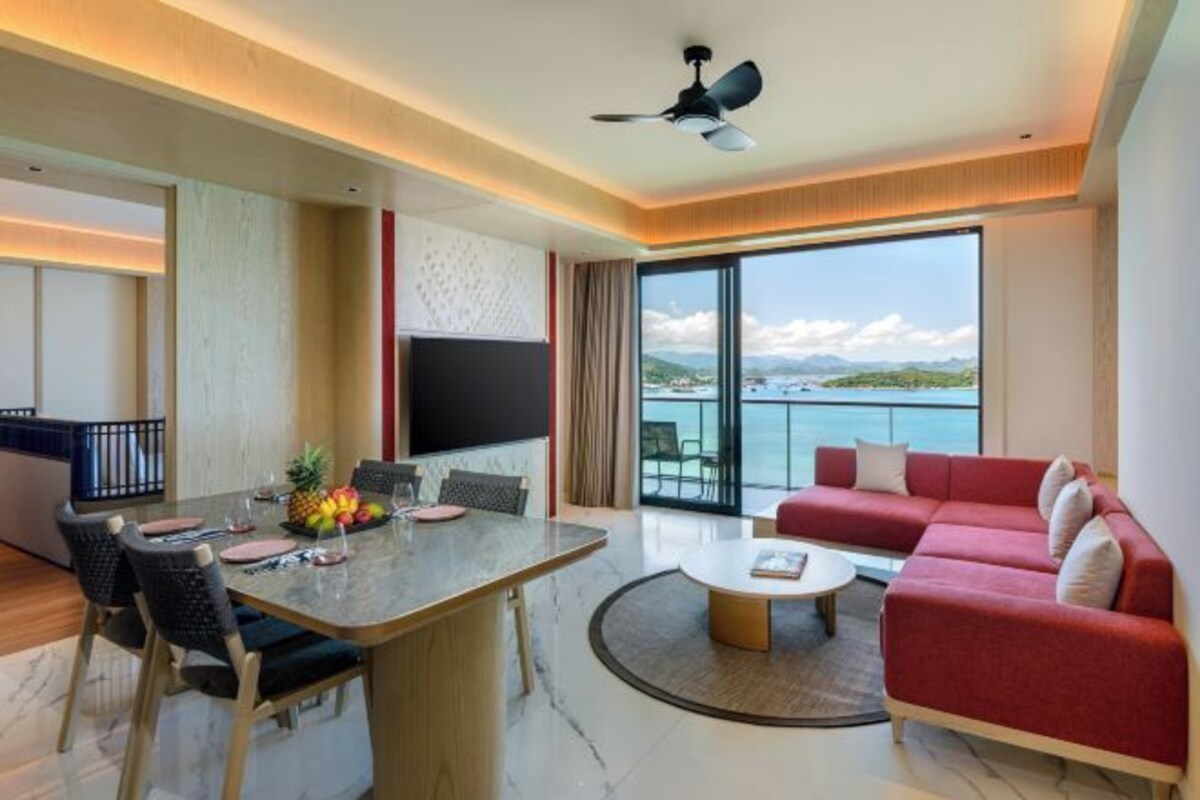A modern hotel room with a dining area, a red sofa, a flat-screen TV, and a balcony with a view of the sea.