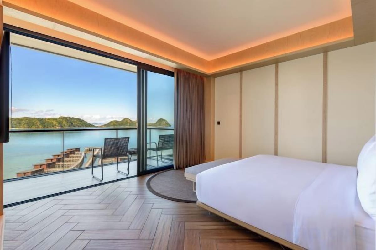 A hotel room with a bed, a view of a body of water, and a balcony with seating.