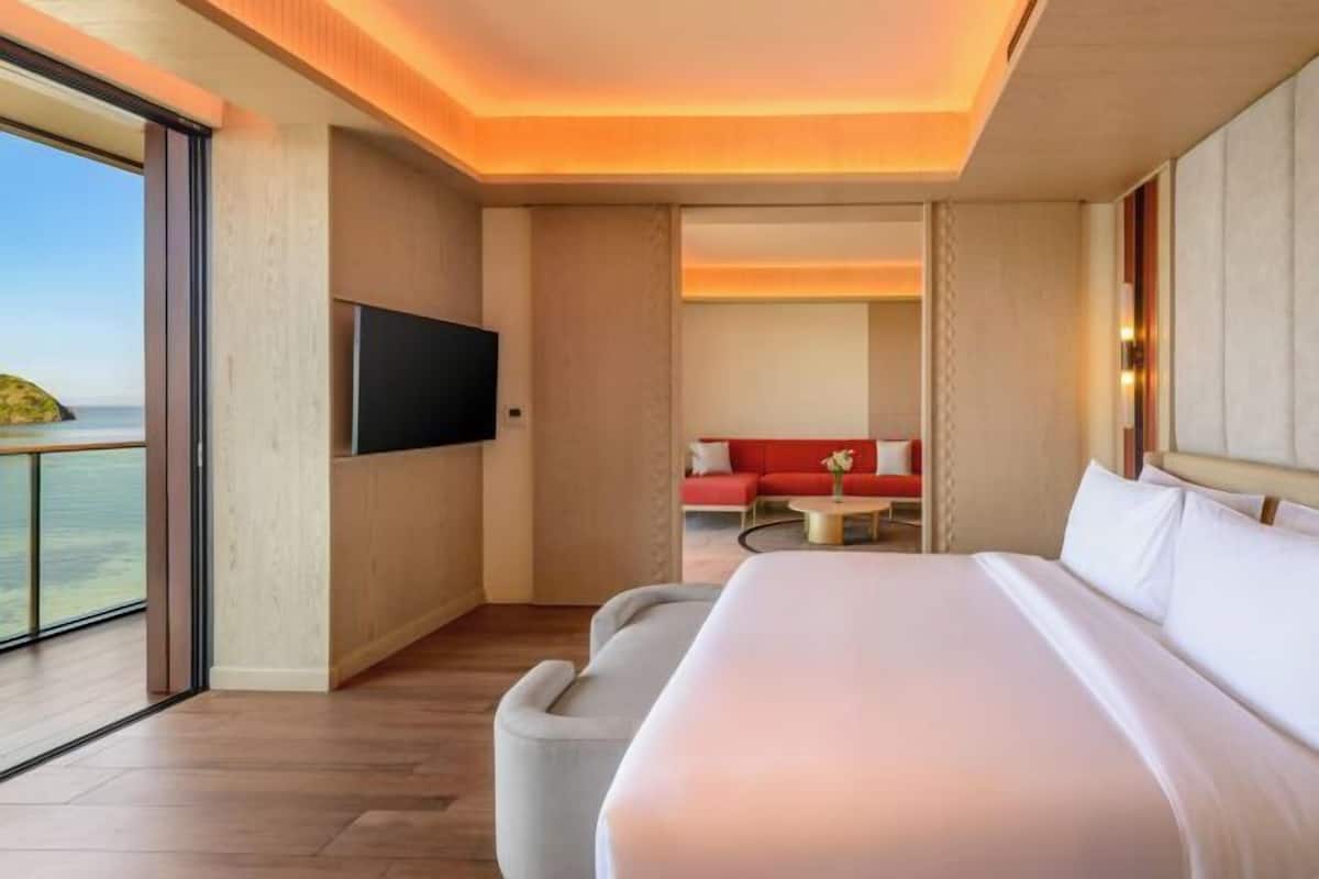 Ein modernes Hotelzimmer mit einem großen Bett, einem Flachbildfernseher, einem Balkon mit Meerblick und einer roten Couch im Hintergrund.