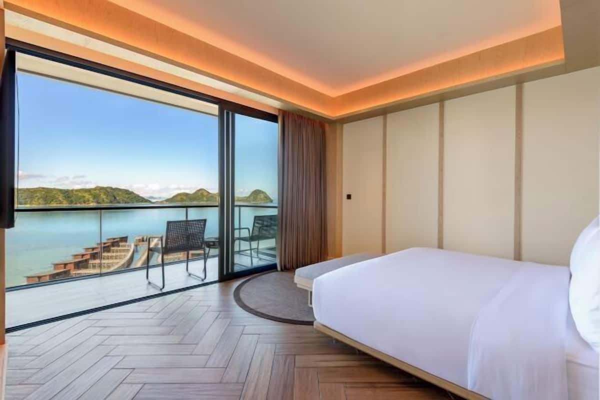 Ein Hotelzimmer mit einer großen Schiebeglasdoor, die auf einen Balkon mit Blick auf ein Gewässer und Hügel führt.