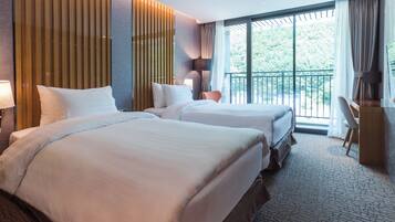 A hotel room with two beds, a desk, a chair, and a balcony with a view of greenery.