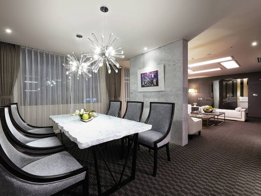 Une chambre d’hôtel moderne dotée d’un coin repas comprenant une table blanche et des chaises grises à revêtement en tissu, un lustre et une grande fenêtre ornée de rideaux légers.