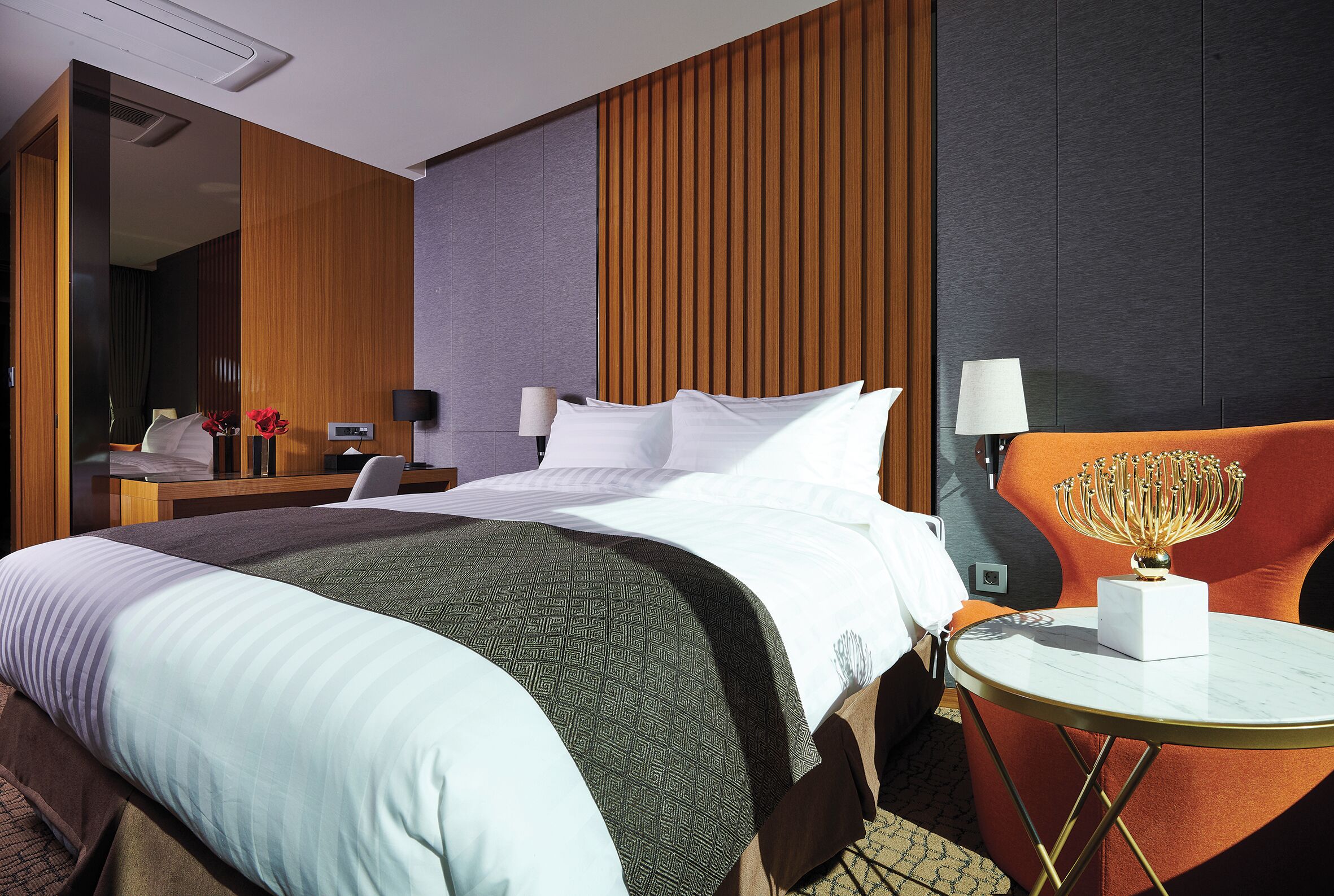 A hotel room with a bed, a desk with a chair, a round table with a lamp, and a decorative item on the table.