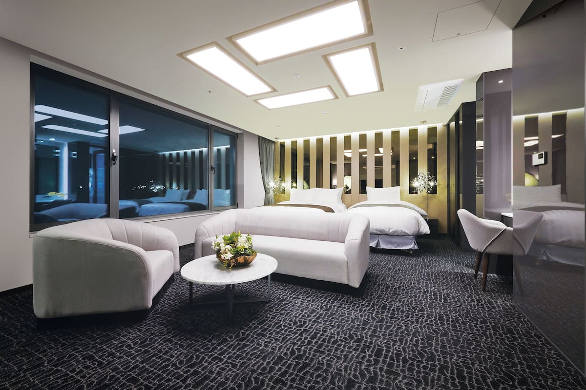 Moderni hotellihuone, jossa on suuri ikkuna, sohva, nojatuoli ja pieni pöytä, jonka päällä on kukkia.