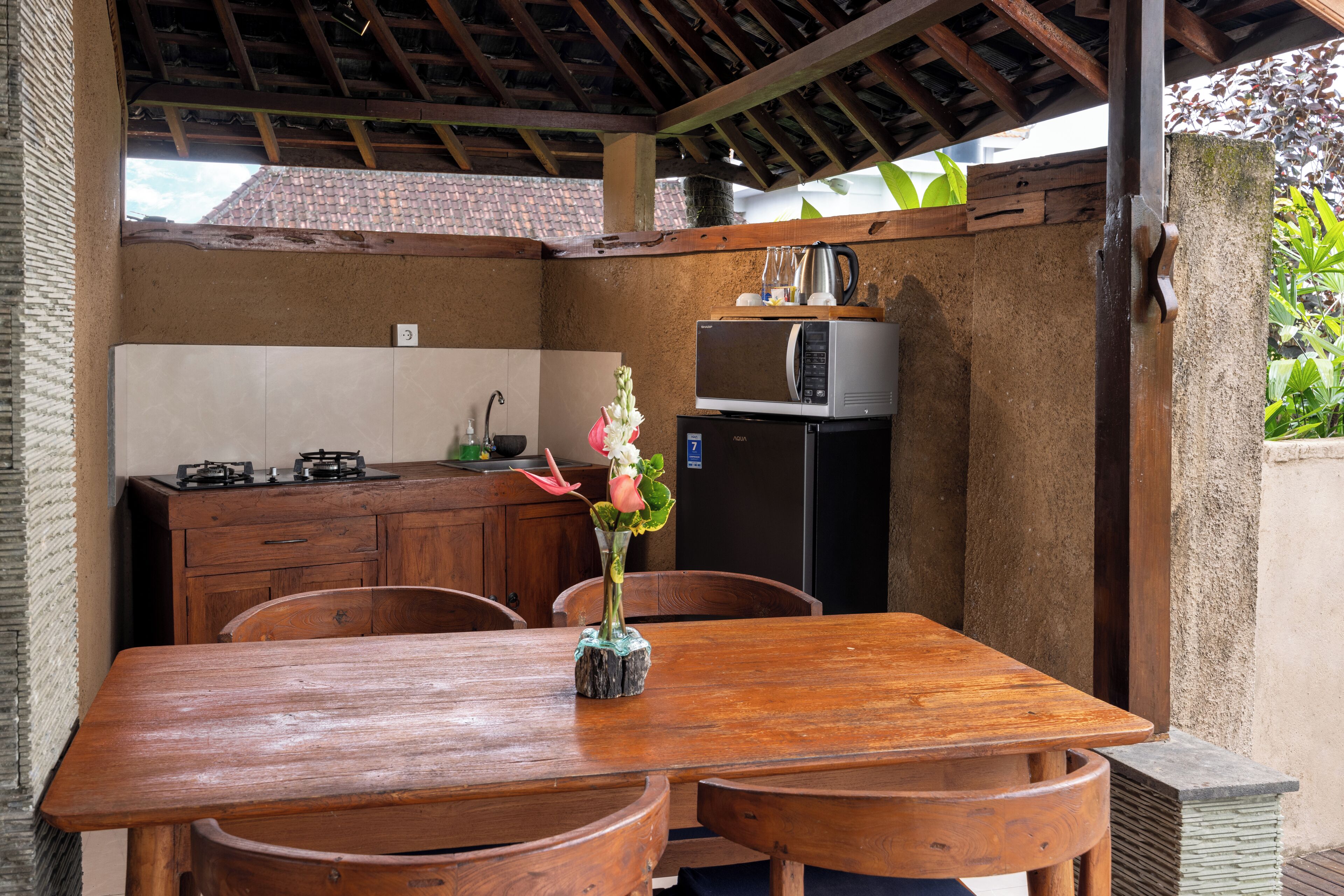A wooden dining table with chairs, a small kitchen area with a microwave and a refrigerator, and a vase with flowers on the table.