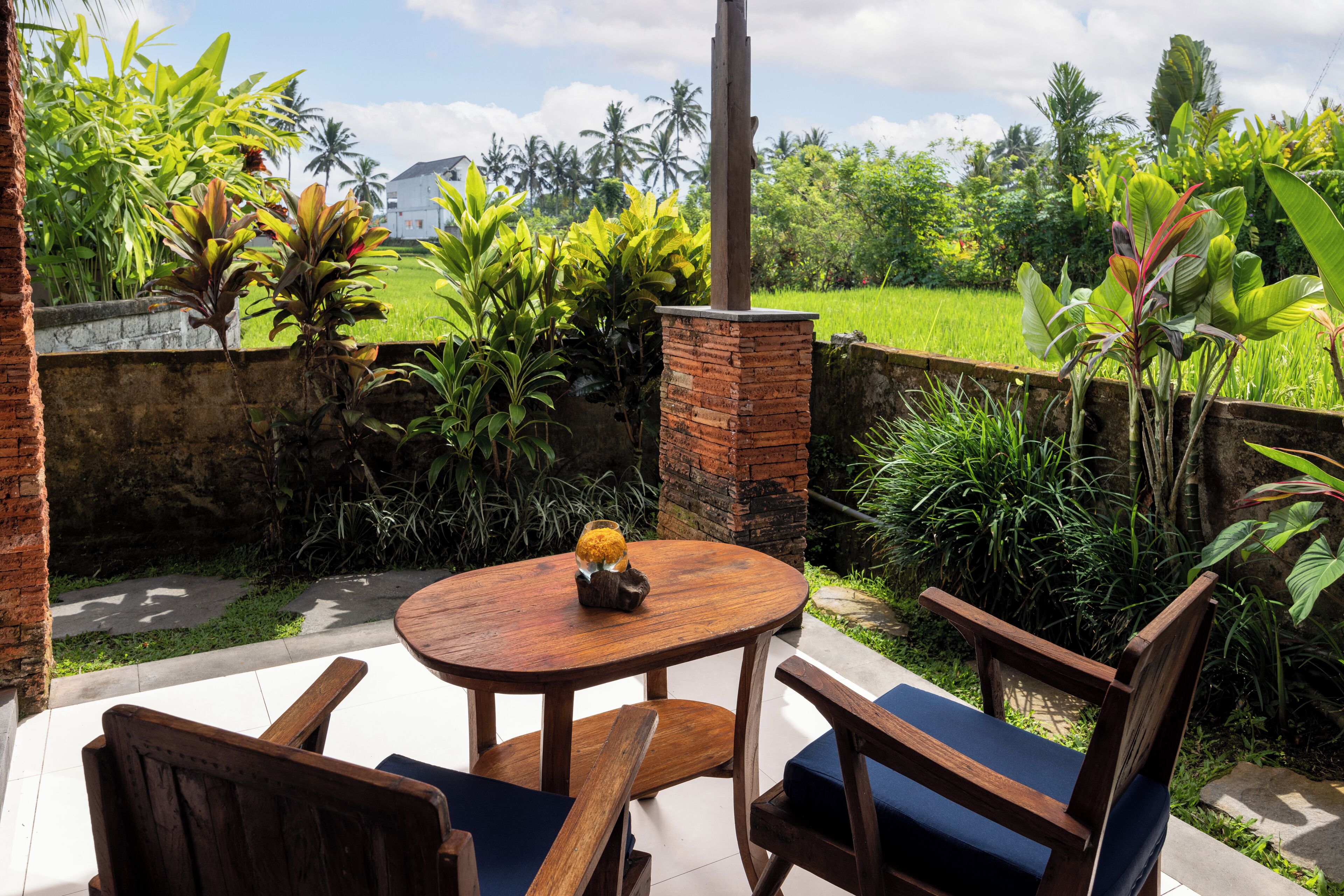 A round wooden table with two chairs, surrounded by lush greenery and a brick wall.
