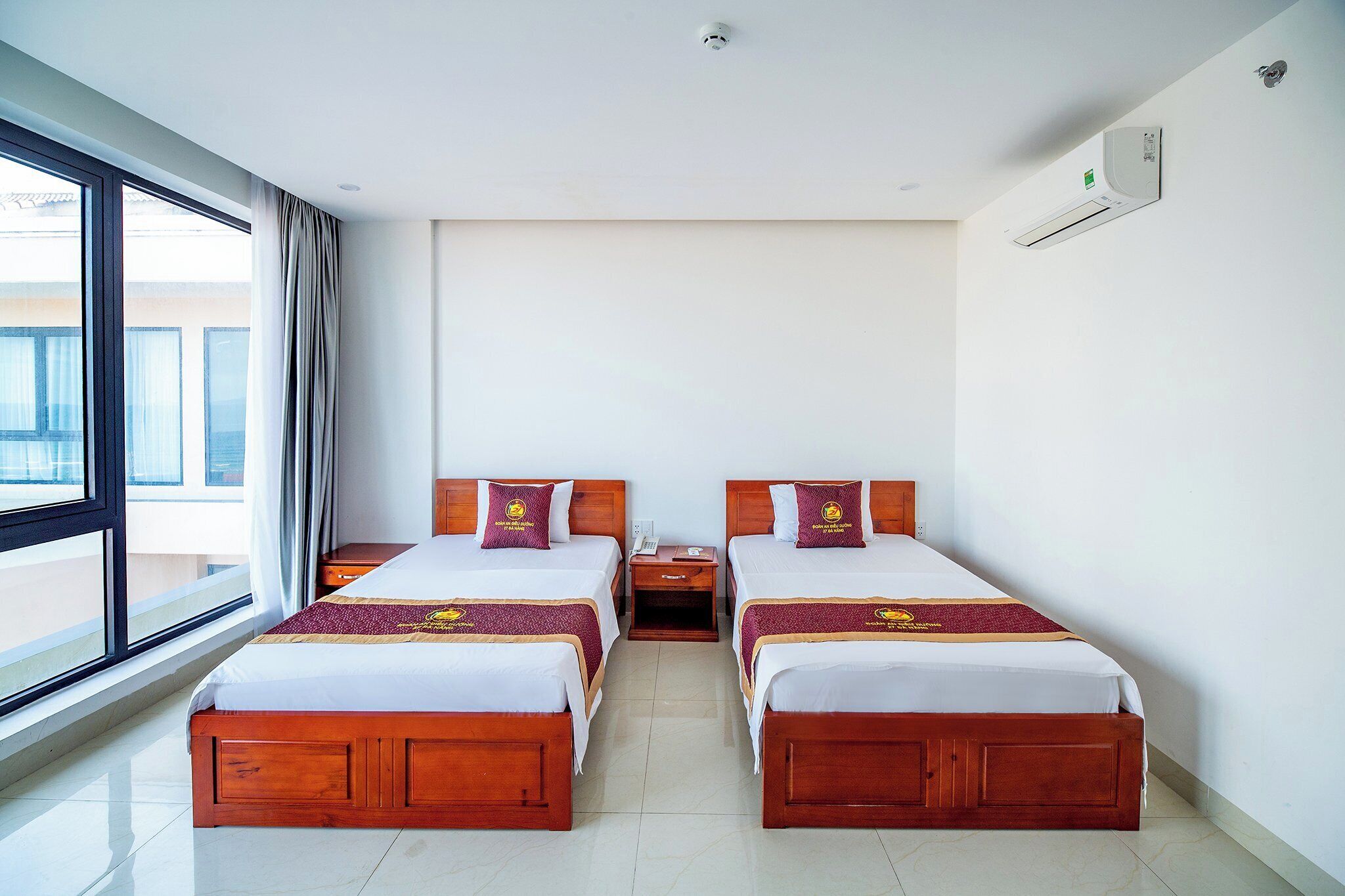 A hotel room with two wooden beds, a nightstand, a window with curtains, and an air conditioning unit on the wall.