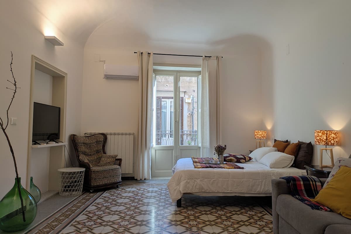 Ein Schlafzimmer mit Bett, Sessel, Sofa, Fernseher und einem Fenster mit Vorhängen.