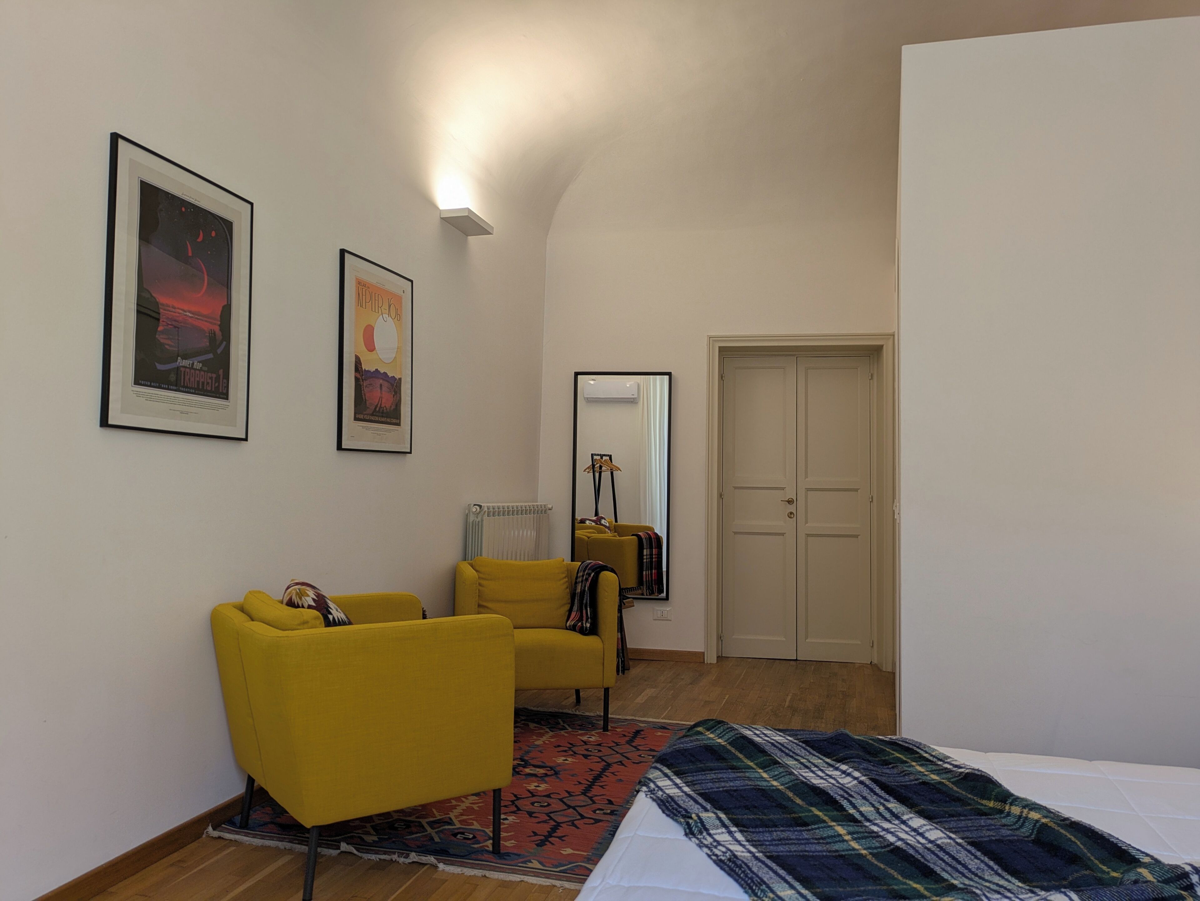 A room with a yellow armchair, a bed with a plaid blanket, a radiator, a mirror, and two framed posters on the wall.