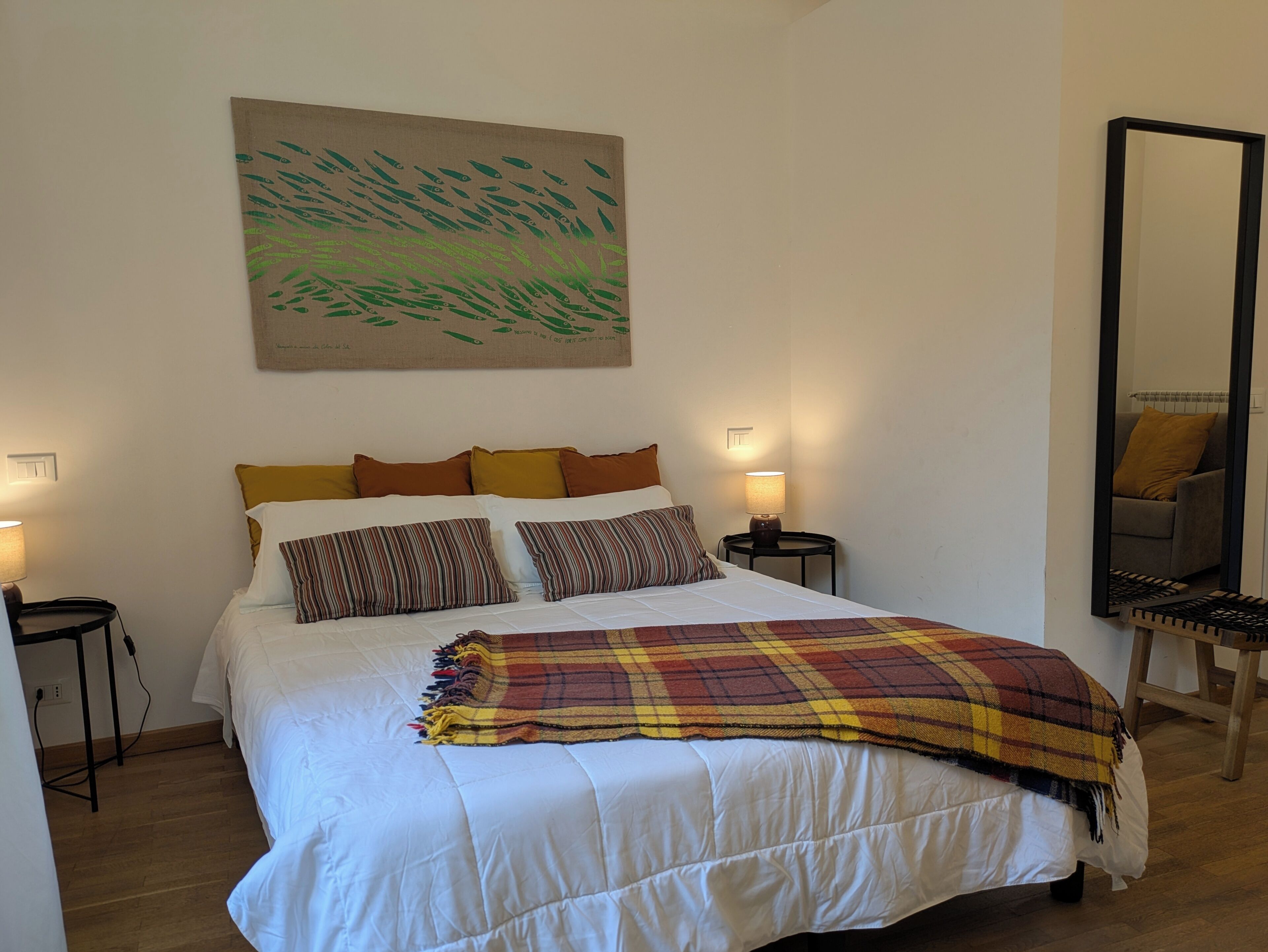 Una cama bien hecha con una colcha escocesa, dos mesitas de noche con lámparas, un cuadro abstracto grande en la pared y un espejo en la pared.