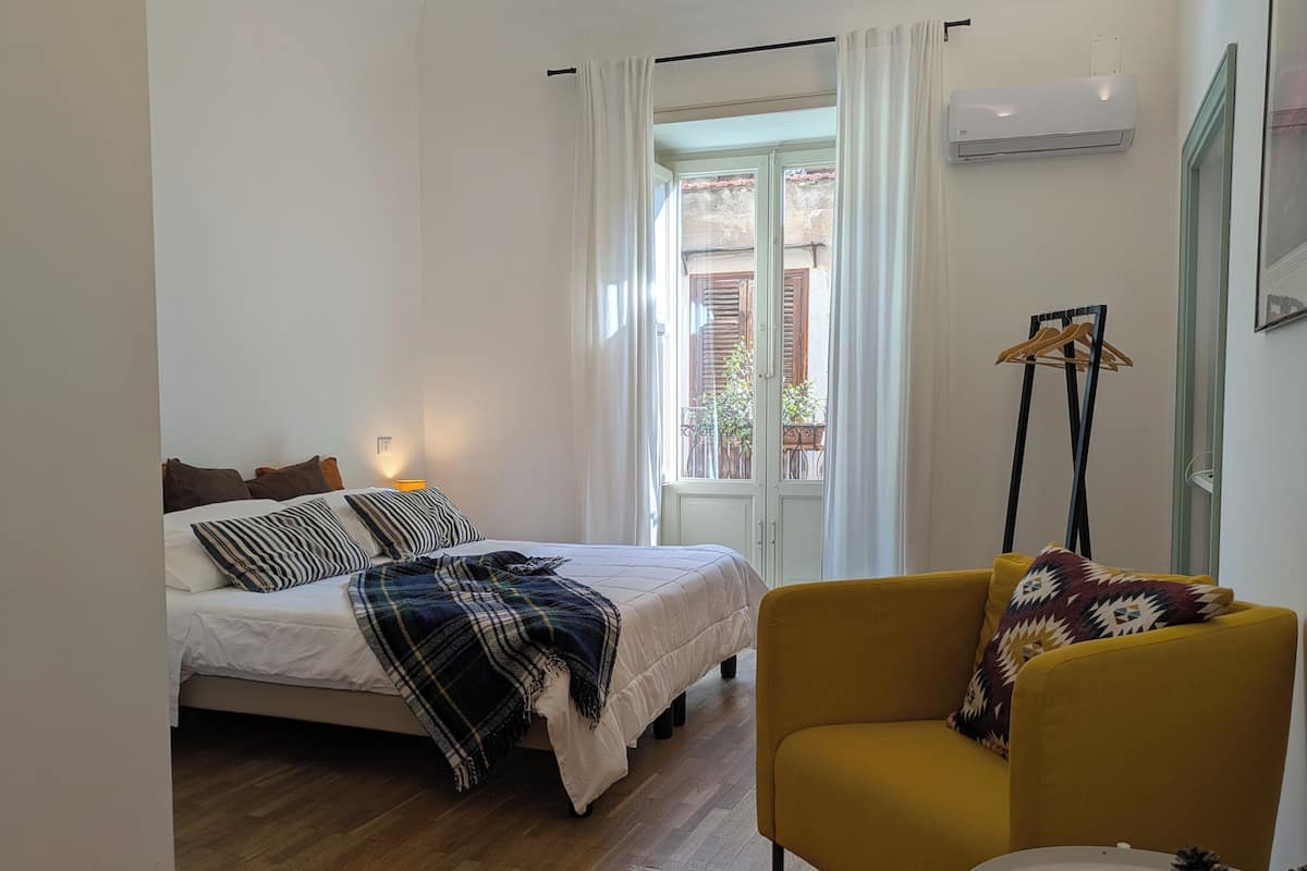 Ein Schlafzimmer mit einem Bett, einem gelben Sessel, einem Fenster mit Vorhängen und einer Klimaanlage.