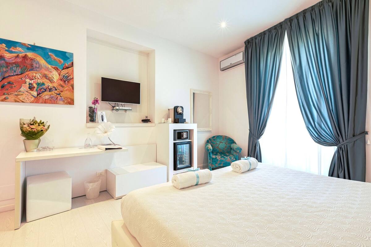 A modern hotel room with a large bed, a desk with a flat-screen TV, a painting on the wall, and a window with curtains.