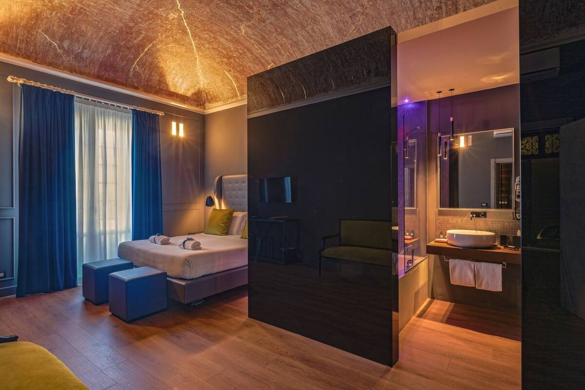 A modern hotel room with a wooden ceiling, a bed with a headboard, a desk, a chair, and a bathroom with a sink and mirror.