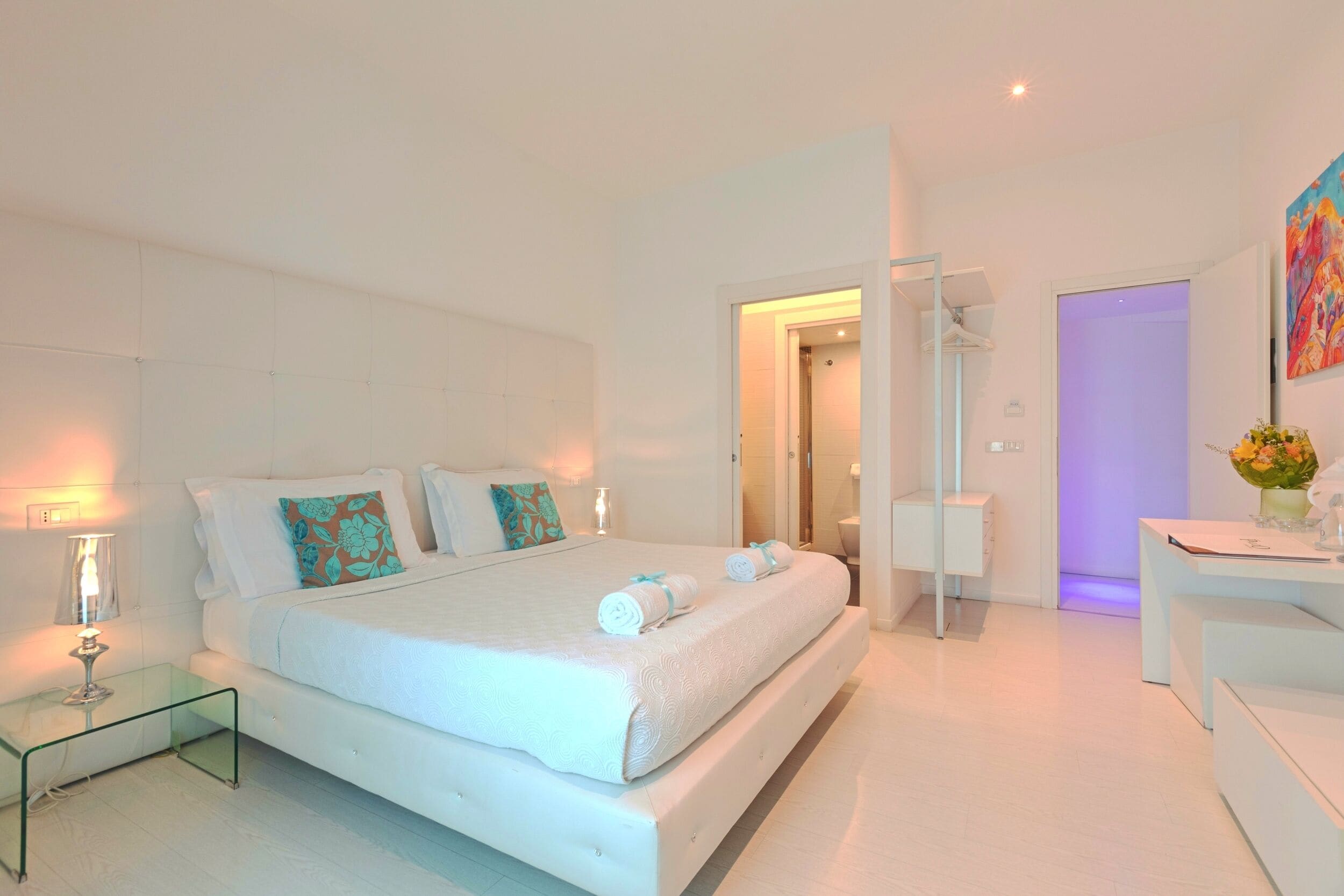 A modern bedroom with a large bed, bedside tables, a desk, and a bathroom visible through an open door.