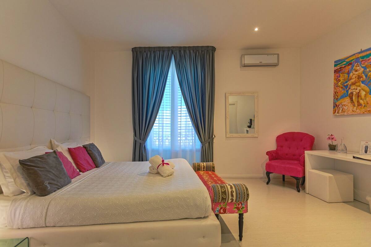 A bedroom with a large bed, a desk, a chair, and a window with curtains.