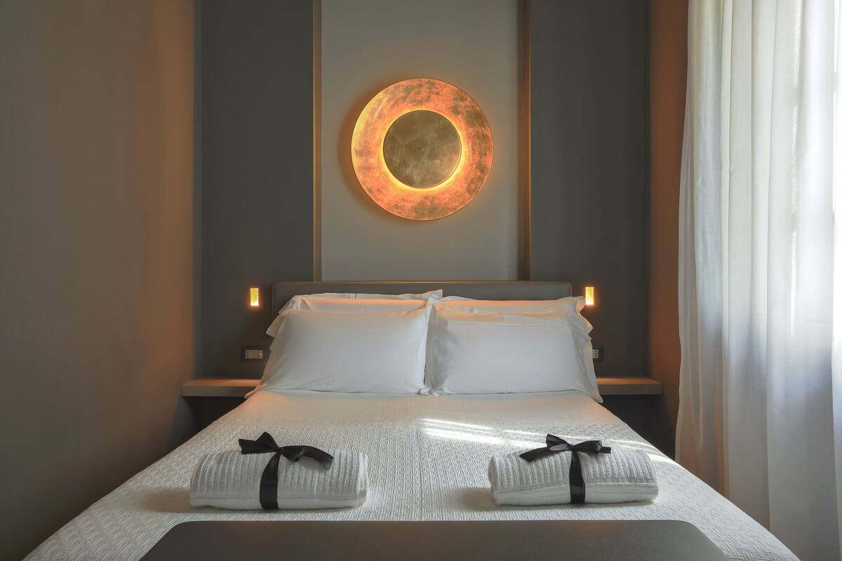 A neatly made bed with white linens and two towels with black ribbons, a circular wall light, and bedside lamps.