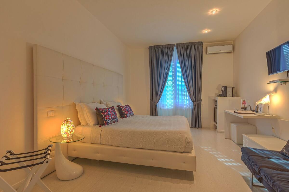 A modern bedroom with a large bed, a bedside table with a lamp, a chair, a desk, and a TV mounted on the wall.