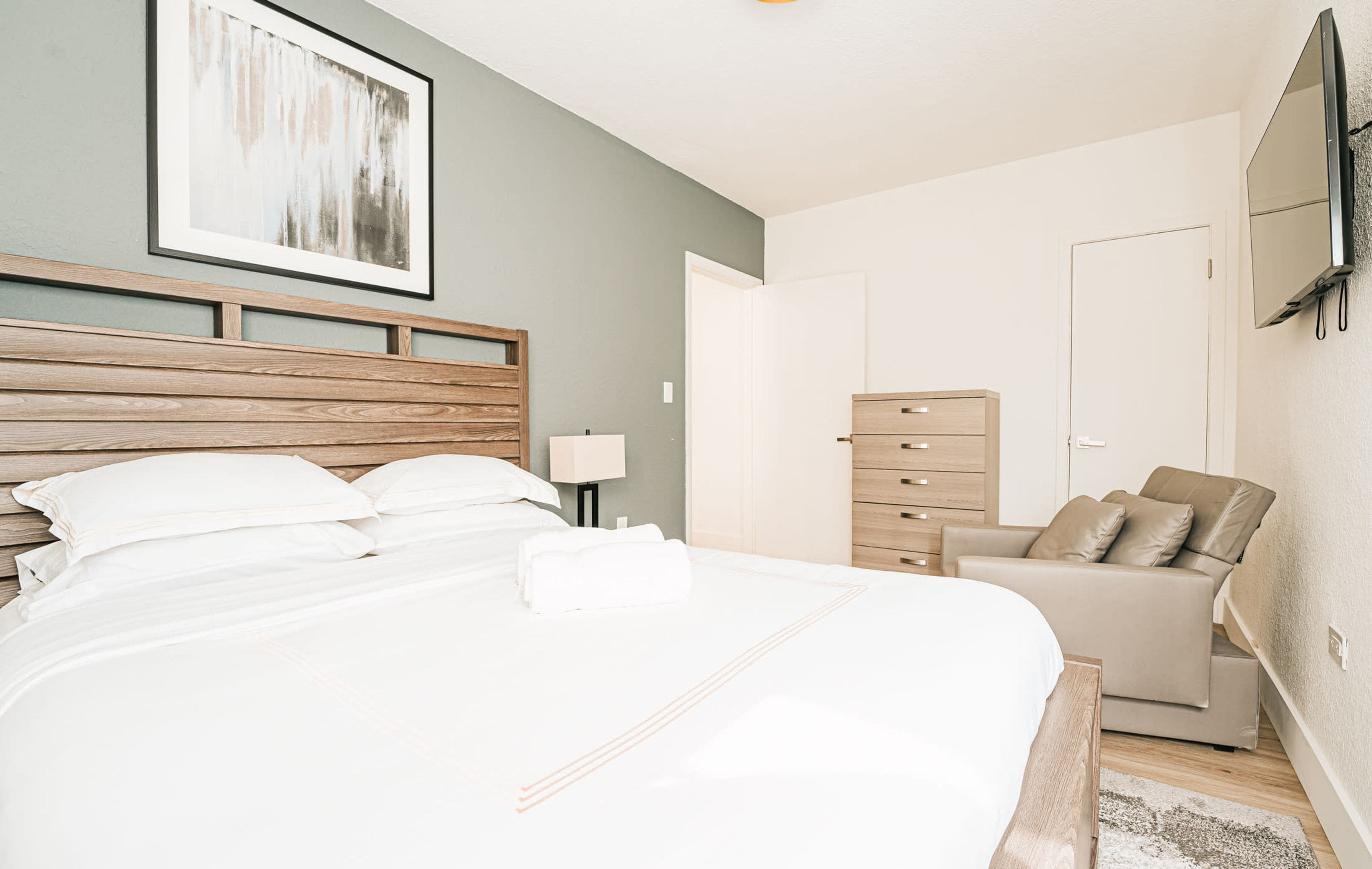 A modern bedroom with a large bed, a wooden headboard, a dresser, a sofa, and a flat-screen TV mounted on the wall.