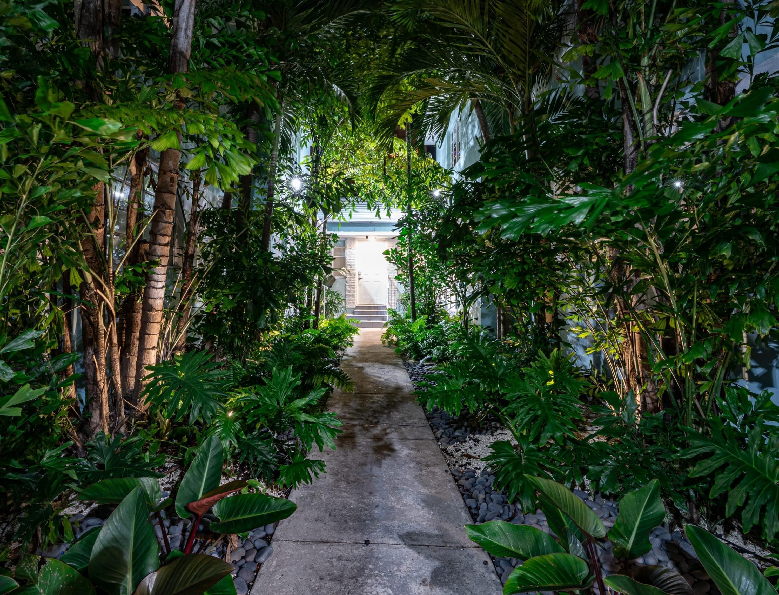 A pathway flanked by lush greenery leading to a building with a white facade and a stone entrance.