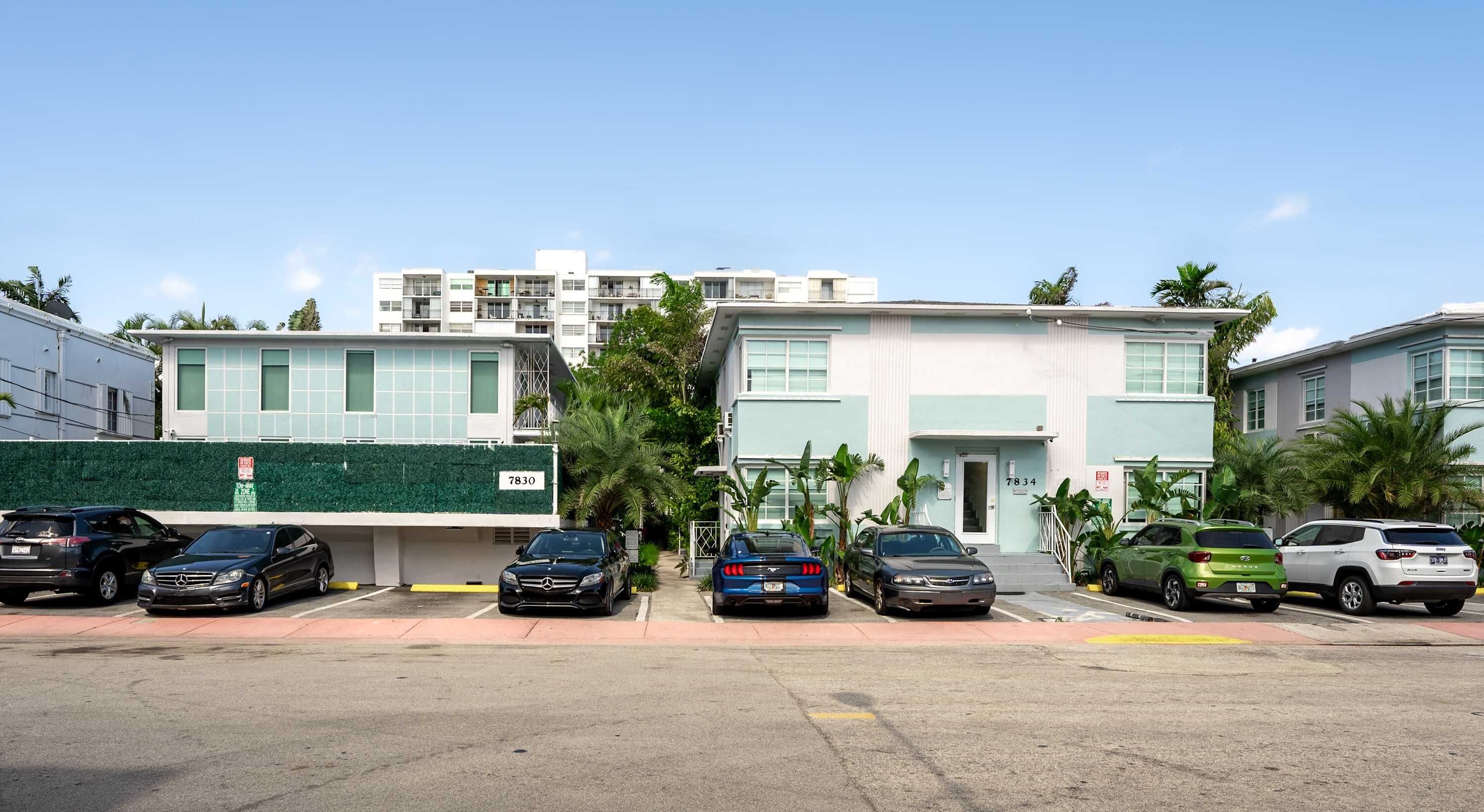 A row of modern two-story buildings with numbered doors, parked cars, and a green hedge.