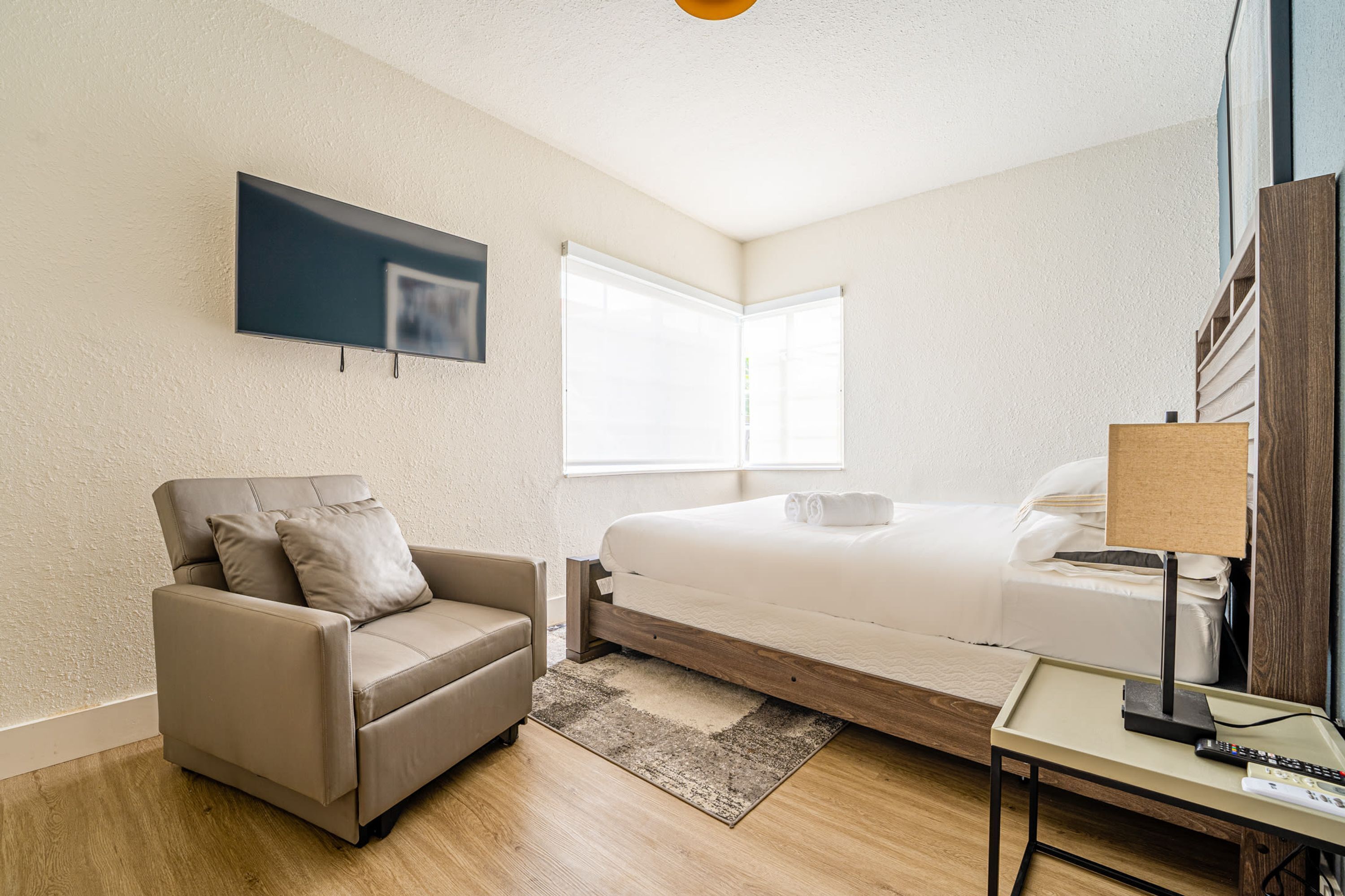 A modern hotel room with a bed, a sofa, a TV mounted on the wall, and a bedside table with a lamp.