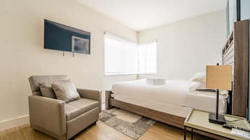 A modern hotel room with a bed, a sofa, a TV mounted on the wall, and a bedside table with a lamp.