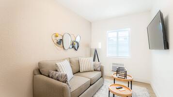 Un salon avec un canapé beige, deux tables d’appoint, une lampe et un téléviseur fixé au mur.