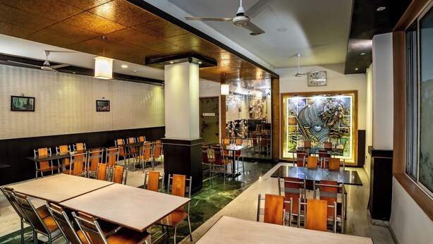 A restaurant with multiple tables and chairs, a ceiling fan, and a decorative wall with a clock. - Hotel RKV Residency (Krishnagiri)